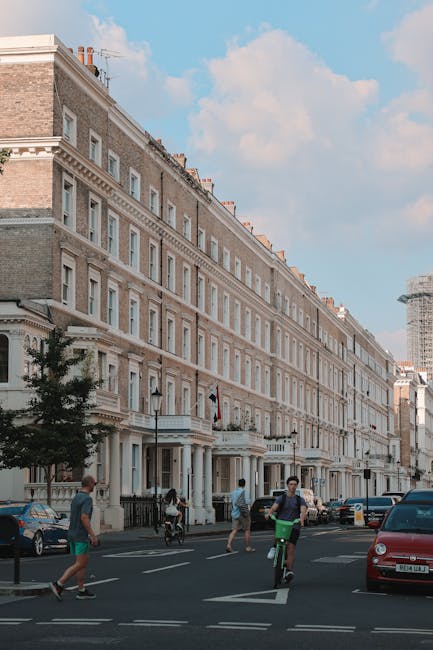 A street scene outside a long, multi-storey Georgian-style building with ornate window frames and a light-colored facade in South Kensington. In the foreground, several people are walking and cycling 