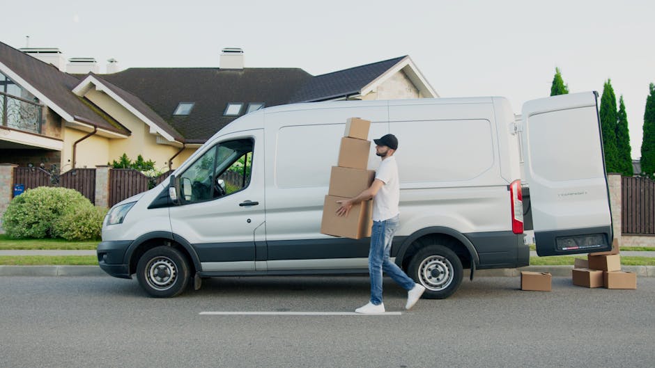 A man wearing casual clothing and a black cap is standing on a suburban street, carrying three stacked cardboard boxes towards a white panel van parked on the roadside. The van has its side door open, revealing an interior prepared for loading, and additional cardboard boxes are seen on the pavement nearby. The residential area features modern houses with pitched roofs, large windows, and fenced gardens with greenery. The scene captures the loading process during a home relocation, with the man actively lifting the boxes and preparing for furniture transport or packing and moving activities. The environment includes street pavement, the house facade, and neatly trimmed bushes along the road, under natural daylight.