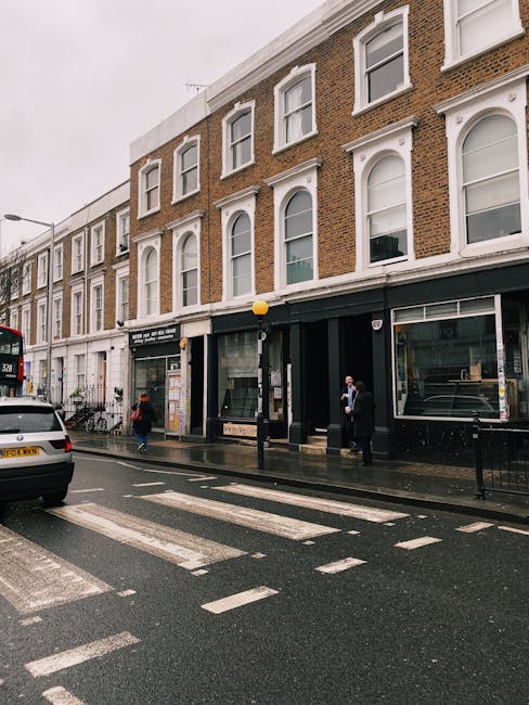 A street scene capturing a three-storey brick building with large arched windows on the upper floors and storefronts at ground level, located on a wet pavement in South Kensington. A black removal van from Removal Van South Kensington is parked outside the building, with its rear doors open for loading or unloading. Several people are visible on the sidewalk, some engaged in conversation while others are preparing items for moving, such as cardboard moving boxes and wrapped furniture. One individual is holding a piece of furniture or a box, possibly assisting with the home relocation process, while another appears to be guiding the loading process. The overall scene suggests an ongoing furniture transport or moving operation, supported by the presence of packing materials, trolleys, and vehicle equipment. Overhead, a yellow spherical streetlight is attached to a black pole, and a double-decker bus is partially visible on the street, emphasizing the urban environment typical of London. The muted lighting indicates overcast weather suited for professional relocation services, aligning with the process of packing and moving house in Kensington and Chelsea.