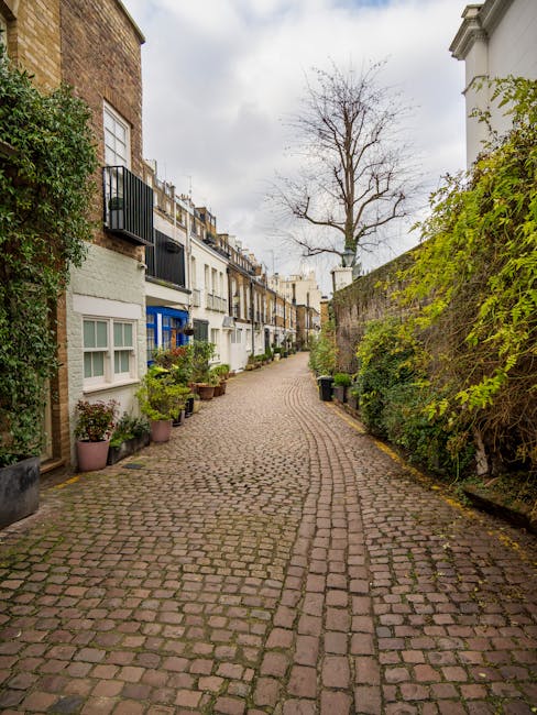A narrow cobbled street in a residential area with a gentle curve, lined with terraced houses featuring white facades, bay windows, and small front gardens adorned with potted plants and greenery. On the right side, there is a tall stone wall partially covered with dense green vines, and a large leafless tree extends above the rooftops. The pavement and street appear damp, indicating recent rain, and the sky is overcast with grey clouds. The scene depicts a quiet urban neighbourhood, suitable for house removals or home relocation services, such as those offered by Removal Van South Kensington, emphasizing the typical setting for furniture transport and moving logistics.