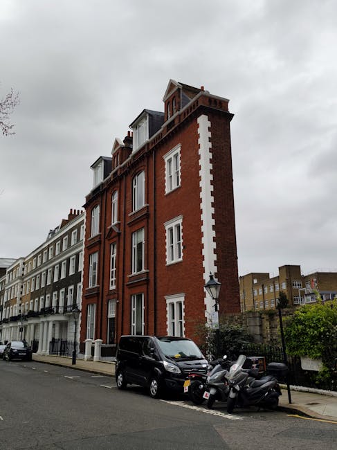 A multi-storey red brick building with white window frames and decorative architectural details, located on a residential street with a grey overcast sky overhead. Parked in front of the building is a black van, along with several motorbikes and scooters on the pavement. The scene suggests a typical urban setting suitable for house removals and furniture transport, with the building's entrance not visible and the street clean and orderly. The image aligns with the context of home relocation services offered by Removal Van South Kensington, emphasizing the urban environment and street-level access for removals in South Kensington.