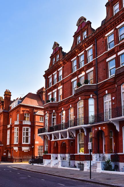 A narrow cobblestone street in South Kensington lined with multi-storey red-brick buildings featuring white window frames and black wrought-iron balconies. On the left, there is a storefront with a black sign displaying 'HEZZO' and a large glass window, with a black street lamp positioned nearby. On the right, several shopfronts with green and black awnings are visible, along with bollards along the pavement. The street appears to be part of a residential and commercial area suitable for house removals and furniture transport, with natural daylight illuminating the scene and no visible vehicles or moving activity present.