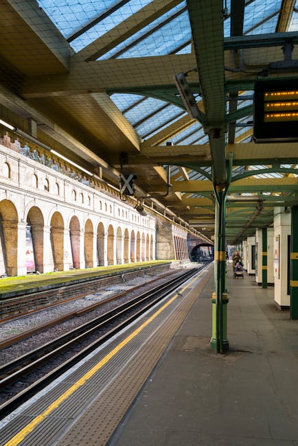 The image shows a railway platform at a station during daytime, featuring train tracks running parallel to the platform. The platform has a grey concrete surface with tactile paving near the edge for safety, and is lined with a green metal canopy supported by vertical poles, providing shelter for waiting passengers. The canopy has a glass roof allowing natural light to illuminate the area, and is equipped with lighting fixtures. Along the platform, there are a few benches, some of which are occupied by commuters, and posters are visible on the walls and pillars. On the right side of the image, a person is waiting with a bicycle, and another individual is walking away from the camera. The station's infrastructure includes a covered walkway at the far end and a tunnel visible in the distance, with a brick wall featuring arched windows on the opposite side of the tracks. This scene is typical of urban rail stations where passengers are preparing for travel, and it aligns with the themes of transportation and home relocation logistics, subtly reflecting services like removals and furniture transport provided by companies such as Removal Van South Kensington.