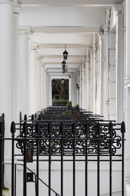 A covered passageway with white-painted columns and ceiling, featuring hanging lantern-style lights. In the foreground, there is a black wrought iron gate with ornate decorative patterns, leading into the corridor. The pathway extends into the distance, where greenery including a large tree and shrubs can be seen through the open end of the passage. The scene is well-lit with natural daylight, and the overall setting appears to be part of a residential or historic property, suitable for house removals or furniture transport services carried out by Removal Van South Kensington as part of their expert home relocation and packing and moving processes.