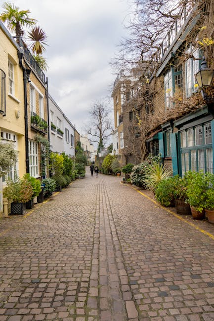 A narrow cobblestone street in South Kensington with residential buildings on both sides, featuring multi-storey houses with large windows, some with balconies and potted plants. The street is lined with various potted plants and small trees, creating a greenery-lined pathway. In the distance, a small group of people are walking away from the camera, with overcast sky overhead. The area appears quiet and residential, with no vehicles in sight, and is suitable for home relocation activities involving tight access. Removal Van South Kensington may utilize this setting for loading or unloading furniture and boxes during a house move, with some objects potentially visible near the buildings or on the pavement.