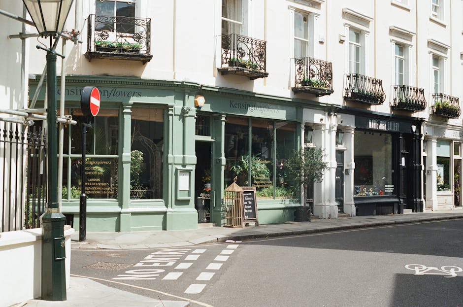 A street scene outside a row of commercial buildings in South Kensington featuring a florist shop with a light green facade and large display windows filled with plants and floral arrangements. Adjacent to it is an art gallery with a black facade and large glass windows. The pavement in front of the shops has a designated loading zone marked by white dashed lines and a small signboard advertising delivery services. A metal pole with a no-entry traffic sign is positioned near the corner, and a bicycle lane symbol is painted on the road. The buildings have multiple stories with ornate wrought-iron balconies and white framed windows, all bathed in natural daylight. This scene showcases typical urban surroundings where house removals or furniture transport may take place, with Removal Van South Kensington often handling complex home relocation tasks in narrow or busy streets.